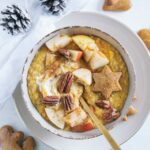 Kürbis-Porridge mit einem Topping aus Apfelstücken, Pekannüssen, einem Lebkuchenstern und Zimt. Goldener Apfel im Porridge. Braune Schüssel auf beigem Teller und weißem Untergrund. Dekoriert mit einem weißen Leinentuch und Zapfen.