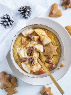 Kürbis-Porridge mit einem Topping aus Apfelstücken, Pekannüssen, einem Lebkuchenstern und Zimt. Goldener Apfel im Porridge. Braune Schüssel auf beigem Teller und weißem Untergrund. Dekoriert mit einem weißen Leinentuch und Zapfen.