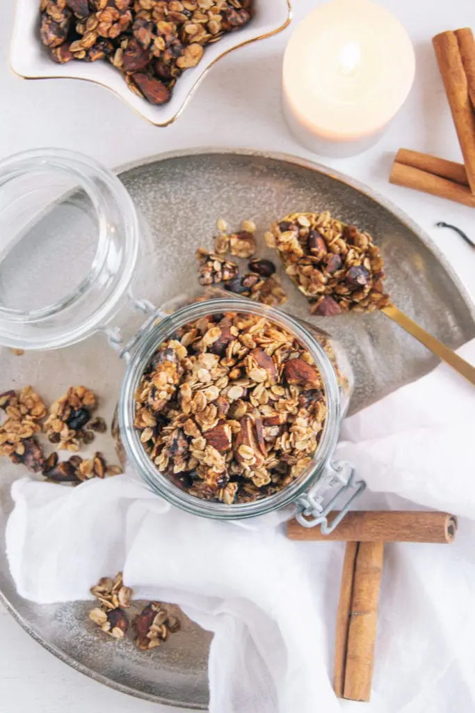 Lebkuchen-Granola in einem Einmachglas mit offenem Deckel auf einem weißen Tuch und einem runden, goldenen Servierteller. Goldener Löffel mit Granola neben dem Glas. Aufnahme von oben.