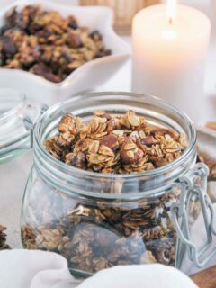 Lebkuchen-Granola in einem Einmachglas mit offenem Deckel. Dekoriert mit einer Kerze und mehr Granola in einer kleinen Sternenschale im Hintergrund. Aufnahme im 45-Grad-Winkel.