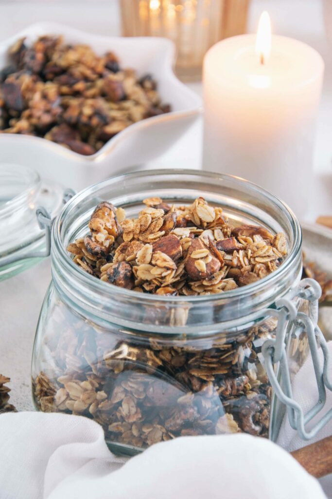 Lebkuchen-Granola in einem Einmachglas mit offenem Deckel. Dekoriert mit einer Kerze und mehr Granola in einer kleinen Sternenschale im Hintergrund. Aufnahme im 45-Grad-Winkel.