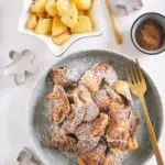 Lebkuchen-Schmarrn auf einem grau-grünen Teller mit goldener Gaben. Daneben Bratapfel-Orangenkompott in einer weißen, sternförmigen Schale und Lebkuchengewürz in einer kleinen Schale. Keksausstecher als Dekoration. Aufnahme von oben auf weißem Untergrund.
