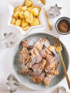 Lebkuchen-Schmarrn auf einem grau-grünen Teller mit goldener Gaben. Daneben Bratapfel-Orangenkompott in einer weißen, sternförmigen Schale und Lebkuchengewürz in einer kleinen Schale. Keksausstecher als Dekoration. Aufnahme von oben auf weißem Untergrund.