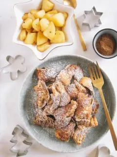 Lebkuchen-Schmarrn auf einem grau-grünen Teller mit goldener Gaben. Daneben Bratapfel-Orangenkompott in einer weißen, sternförmigen Schale und Lebkuchengewürz in einer kleinen Schale. Keksausstecher als Dekoration. Aufnahme von oben auf weißem Untergrund.