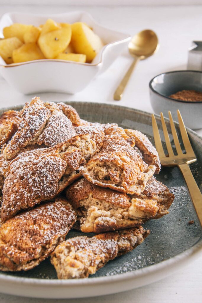 Lebkuchen-Schmarrn auf einem graugrünen Teller mit goldener Gaben. Aufnahme im 45-Grad-Winkel. Bratapfel-Orangenkompott im Hintergrund.