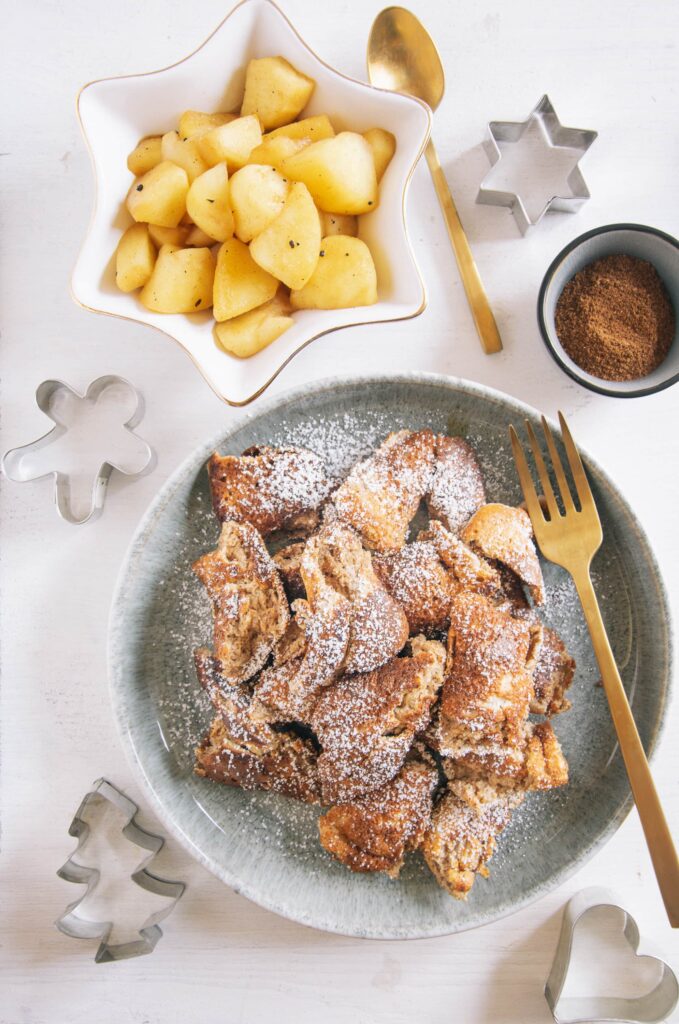 Lebkuchen-Schmarrn auf einem grau-grünen Teller mit goldener Gaben. Daneben Bratapfel-Orangenkompott in einer weißen, sternförmigen Schale und Lebkuchengewürz in einer kleinen Schale. Keksausstecher als Dekoration. Aufnahme von oben auf weißem Untergrund.