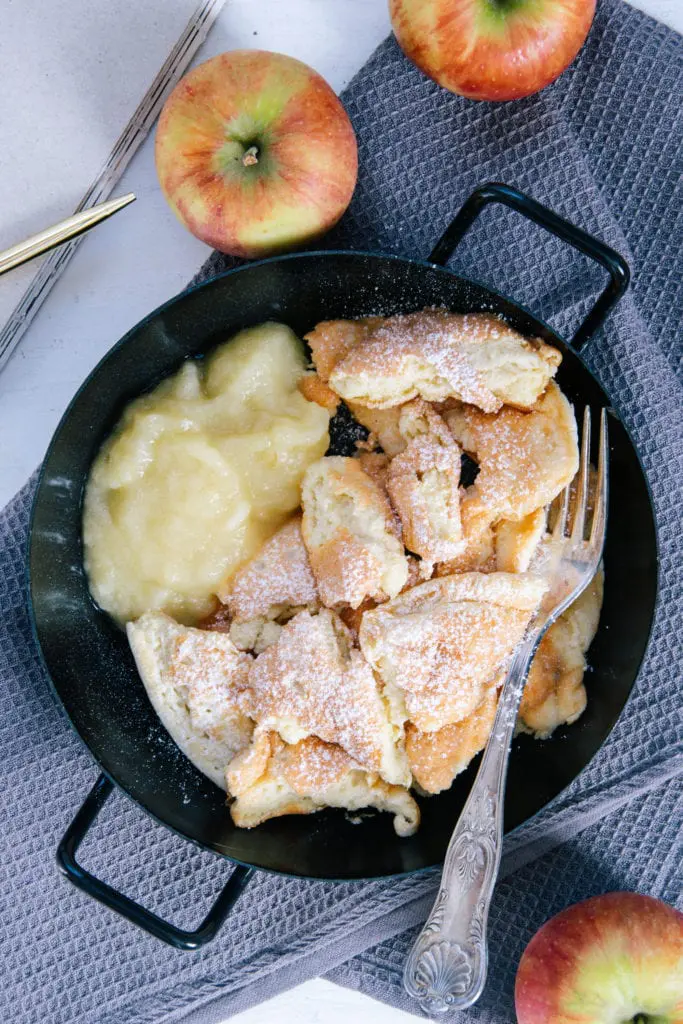 Protein-Kaiserschmarrn mit Apfelmus in einer schwarzen Servierpfanne auf einem grauen Leinentuch. Äpfel als Dekoration. Von oben fotografiert.