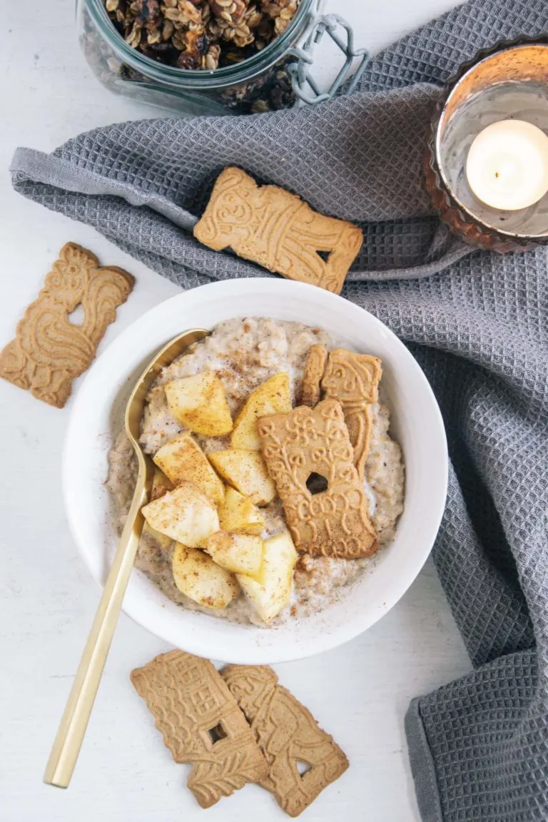 Spekulatius-Porridge mit Bratapfel in einer weißen Schüssel mit goldenem Löffel. Aufnahme von oben. Graues Leinentuch, Kerze und weitere Spekulatiuskekse als Dekoration.