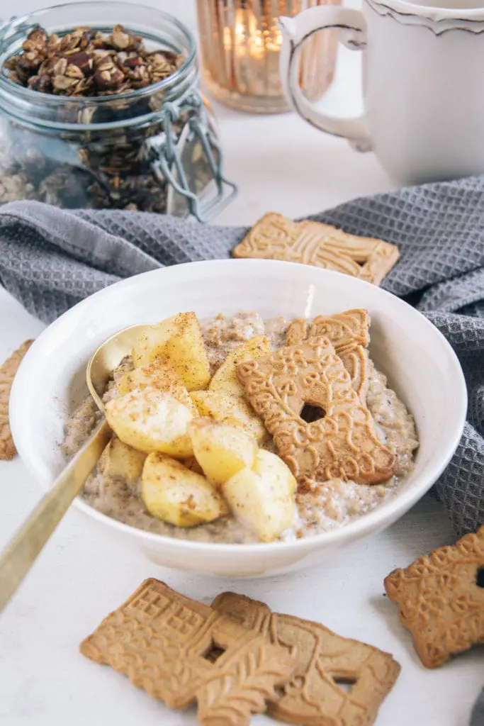 Spekulatius-Porridge mit Bratapfel in einer weißen Schüssel mit goldenem Löffel. Aufnahme im 45-Grad-Winkel.