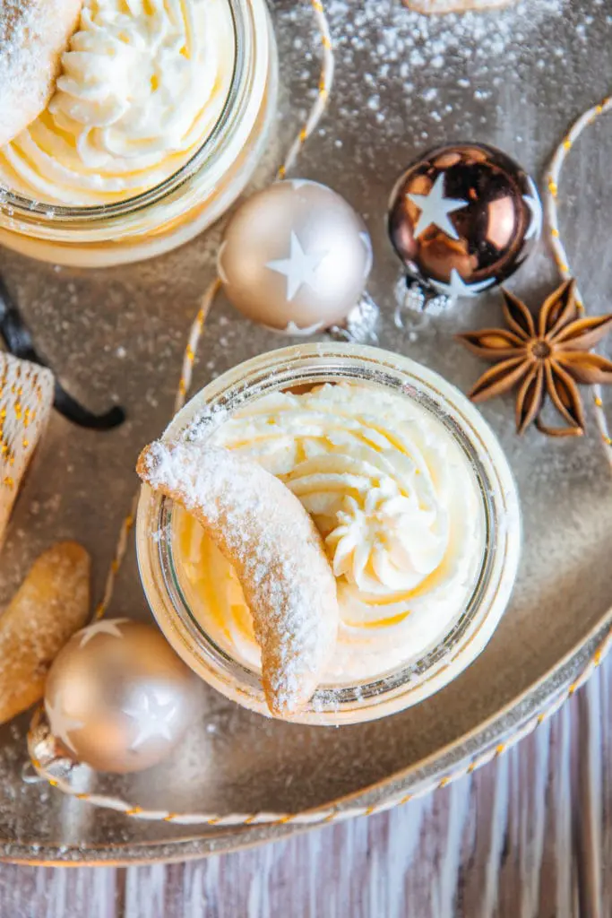 Nahaufnahme von oben von Vanillekipferl-Cheesecake-Dessert mit Mandelnougat. Die Sahne im Glas und das Vanillekipferl als Topping sind detailliert sichtbar, daneben zwei kleine Christbaumkugeln mit Sternen und ein Anisstern.