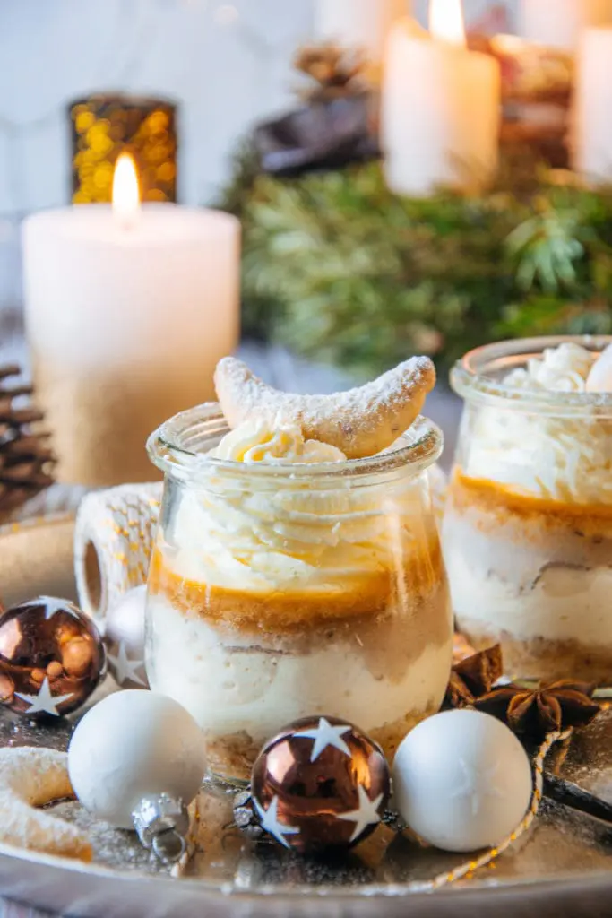 Zwei Gläser Vanillekipferl-Cheesecake-Dessert mit Mandelnougat im kleinen Tulpenglas fast bildfüllend platziert und knapp über Augenhöhe fotografiert. Im Hintergrund brennende Kerzen vom Adventkranz. Das Bild strahlt Wärme und Gemütlichkeit aus.