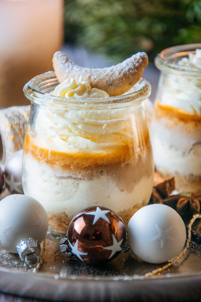 Vanillekipferl-Cheesecake-Dessert mit Mandelnougat im kleinen Tulpenglas fast bildfüllend platziert und knapp über Augenhöhe platziert. Im Hintergrund oben grüner Akzent durch einen Adventkranz.