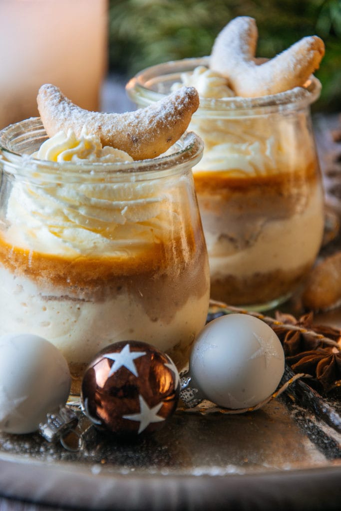 Zwei Gläser Vanillekipferl-Cheesecake-Dessert mit Mandelnougat auf einem goldenen Teller, dekoriert mit kleinen Christbaumkugeln. Fotografiert leicht über Augenhöhe bei warmem, gemütlichem Licht.