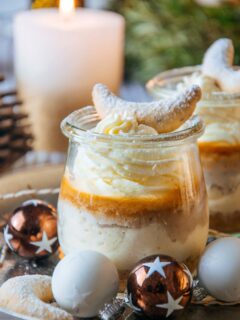 Vanillekipferl-Cheesecake-Dessert mit Mandelnougat in einem kleinen Tulpenglas mit dressierter Sahne und einem Vanillekipferl als Topping. Aufnahme fast auf Augenhöhe, sodass die Schichten im Glas gut erkennbar sind. Brennende Kerze im Hintergrund.