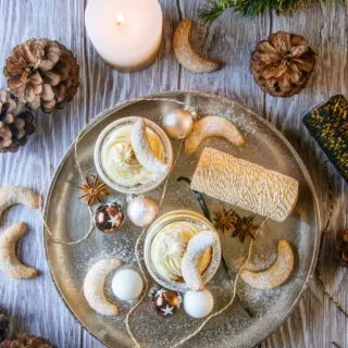 Zwei Gläser Vanillekipferl-Cheesecake-Dessert mit Mandelnougat auf einem großen goldenen Teller, verziert mit Vanillekipferln, kleinen Christbaumkugeln, Glitzergarn und Anissternen. Aufnahme von oben auf braunem Untergrund im rustikalen Stil.