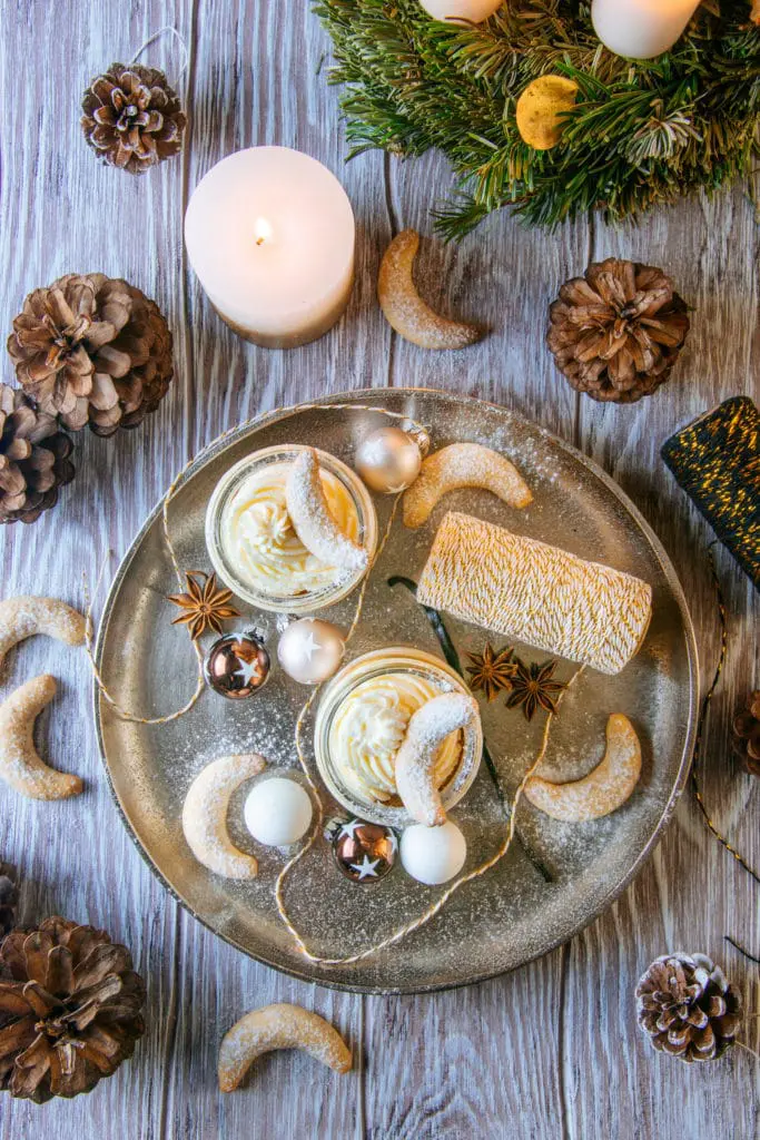 Zwei Gläser Vanillekipferl-Cheesecake-Dessert mit Mandelnougat auf einem großen goldenen Teller, verziert mit Vanillekipferln, kleinen Christbaumkugeln, Glitzergarn und Anissternen. Aufnahme von oben auf braunem Untergrund im rustikalen Stil.