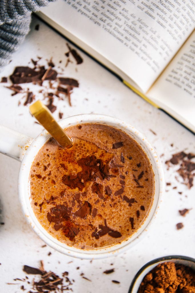 Eine Tasse Kakao auf hellem Untergrund mit Kakaopulver darauf. Goldener Löffel in der Tasse, aufgeschlagenes Buch daneben. Auf dem Kakao befinden sich geschmolzene Schokolade, Zimt und Kokosöl. Nahaufnahme von oben.