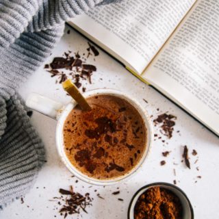 Eine Tasse Kakao von oben fotografiert auf hellem Untergrund mit Kakaopulver darauf. Goldener Löffel in der Tasse, aufgeschlagenes Buch und grauer Pullover daneben.