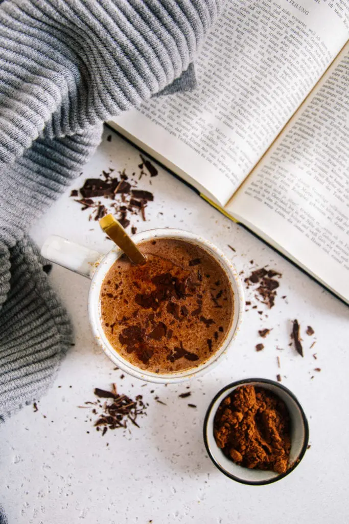 Eine Tasse Kakao von oben fotografiert auf hellem Untergrund mit Kakaopulver darauf. Goldener Löffel in der Tasse, aufgeschlagenes Buch und grauer Pullover daneben.