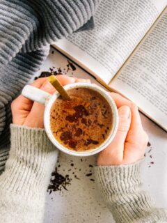 Zwei Hände umschließen eine Tasse Kakao. Aufnahme von oben mit aufgeschlagenem Buch daneben. Warme Farben und hyggeliger Stil.