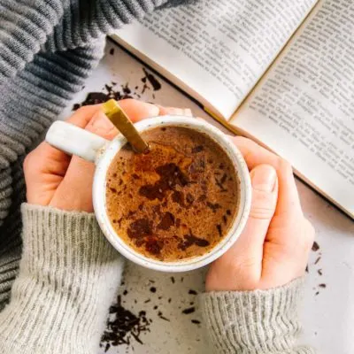 Zwei Hände umschließen eine Tasse Kakao. Aufnahme von oben mit aufgeschlagenem Buch daneben. Warme Farben und hyggeliger Stil.