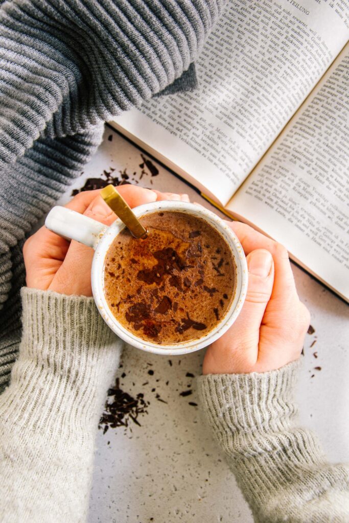 Zwei Hände umschließen eine Tasse Kakao. Aufnahme von oben mit aufgeschlagenem Buch daneben. Warme Farben und hyggeliger Stil.