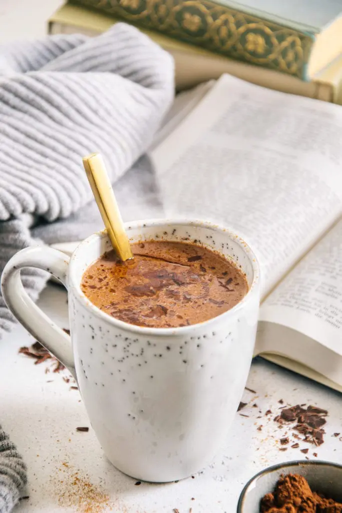 Kakao mit geschmolzener Schokolade und Kokosöl an der Oberfläche in einer graublauen Steingut-Tasse mit goldenem Löffel darin, aufgenommen im 45-Grad-Winnkel mit einem aufgeschlagenen Buch und einem grauen Pullover im Hintergrund