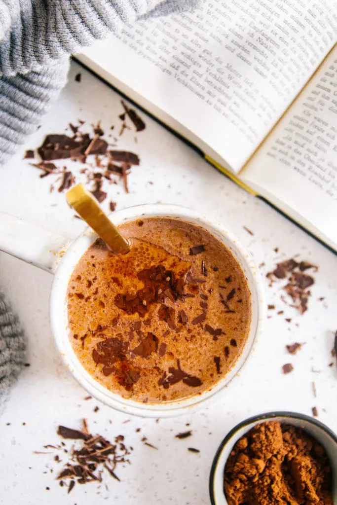 Eine Tasse Kakao von oben fotografiert auf hellem Untergrund mit Kakaopulver darauf. Goldener Löffel in der Tasse, aufgeschlagenes Buch in der rechten oberen Ecke des Bildes.