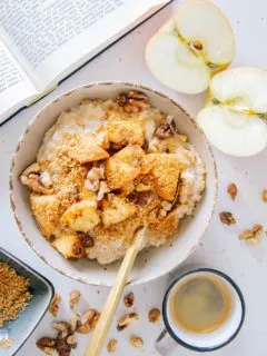 Apfelstrudel-Porridge in einer grauen Schüssel auf hellem Untergrund. Eine Tasse Espresso, ein halbierter Apfel, Walnüsse und ein Buch daneben. Aufnahme von oben.