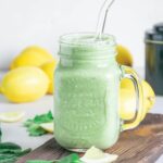 Ein Glas Grüner Smoothie für die Darmgesundheit auf einem rustikalen Brett. Set verziert mit Spinatblättern, Zitronenstücken und Zitronen im Hintergrund. Glasstrohhalm im Smoothie.