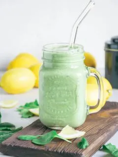 Ein Glas Grüner Smoothie für die Darmgesundheit auf einem rustikalen Brett. Set verziert mit Spinatblättern, Zitronenstücken und Zitronen im Hintergrund. Glasstrohhalm im Smoothie.