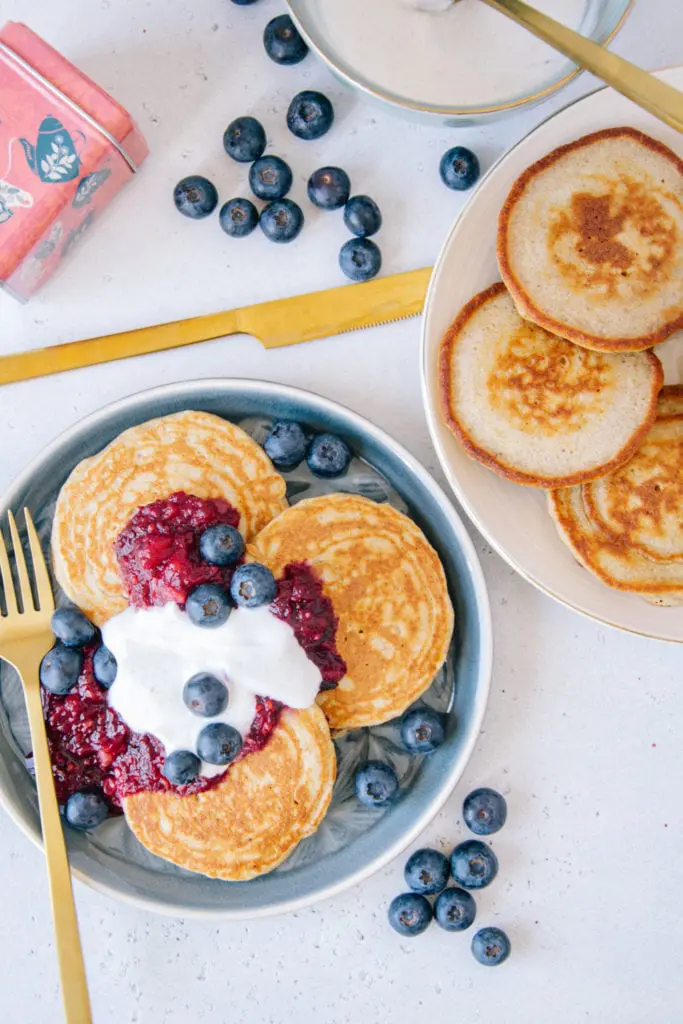 Gesunde glutenfreie Hafer-Chai-Pancakes auf zwei Tellern von oben fotografiert. Ein Teller von rechts angeschnitten. Goldenes Besteck und Heidelbeeren als Verzierung.