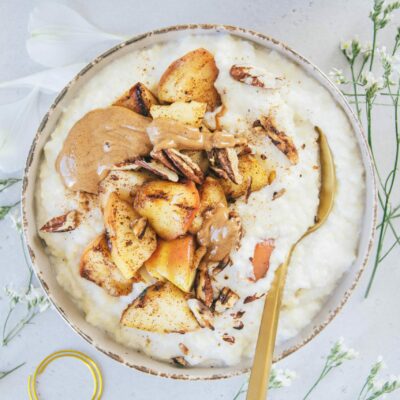 Eine Schüssel Hirse-Porridge mit Bratapfel von oben fotografiert auf hellem Untergrund. Topping aus Bratapfel, Mandelmus und gerösteten Pekannüssen.
