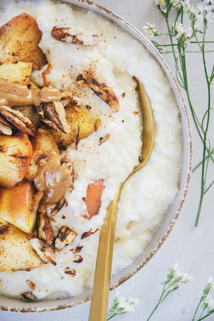 Nahaufnahme von Hirse-Porridge mit Bratapfel von oben. Schüssel von links angeschnitten. Ein goldener Löffel steckt seitlich im Porridge, sodass man sieht, wie cremig es ist.