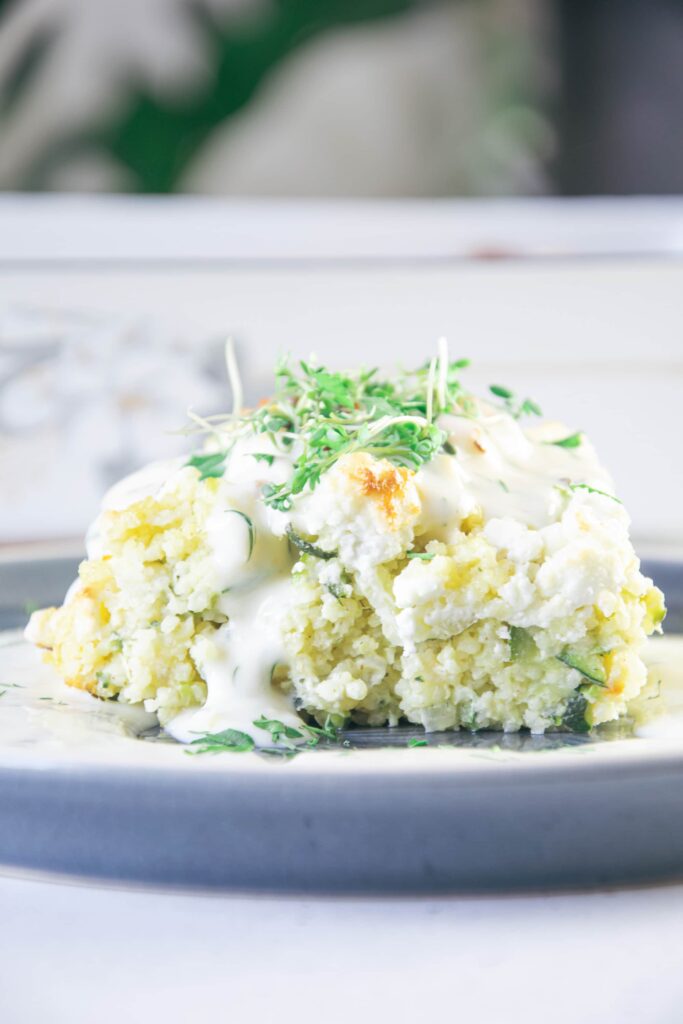 Nahaufnahme von einem Stück Hirse-Zucchini-Auflauf mit Dillsauce auf Augenhöhe. Die Sauce fließt am Auflauf herunter. Der Auflauf ist mit Kresse garniert.