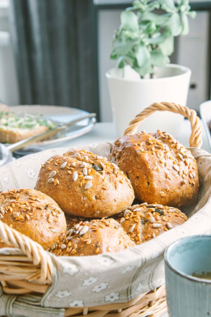 Geflochtener Brotkorb mit frischen Dinkel-Körnerweckerln. Aufnahme im 45-Grad-Winkel. Eine Grünpflanze und ein Teller mit einem aufgeschnittenen Weckerl im Hintergrund.