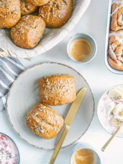 Zwei Dinkel-Körnerweckerl auf einem grauen Teller mit einem goldenen Messer. Zwei Müslischüsseln, zwei Espressotassen, ein Brotkorb und eine Auflaufform mit Zimtschnecken am Bildrand angeschnitten rundherum. Aufnahme von oben.