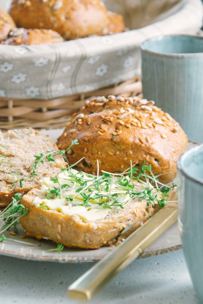 Aufgeschnittenes und mit Butter bestrichenes Dinkel-Körnerweckerl in der Aufnahme auf Augenhöhe. Mit Kresse garniert. Ein weiteres Weckerl im Hintergrund.