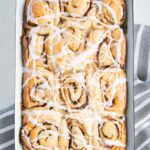Gesündere Zimtschnecken mit Frischkäseglasur in einer alten weißen Emailleform. Aufnahme von oben auf hellem Untergrund. Grau-weiß gestreiftes Küchentuch unter der Form.