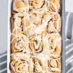 Gesündere Zimtschnecken mit Frischkäseglasur in einer alten weißen Emailleform. Aufnahme von oben auf hellem Untergrund. Grau-weiß gestreiftes Küchentuch unter der Form.