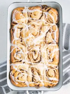 Gesündere Zimtschnecken mit Frischkäseglasur in einer alten weißen Emailleform. Aufnahme von oben auf hellem Untergrund. Grau-weiß gestreiftes Küchentuch unter der Form.