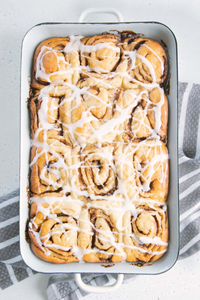 Gesündere Zimtschnecken mit Frischkäseglasur in einer alten weißen Emailleform. Aufnahme von oben auf hellem Untergrund. Grau-weiß gestreiftes Küchentuch unter der Form.