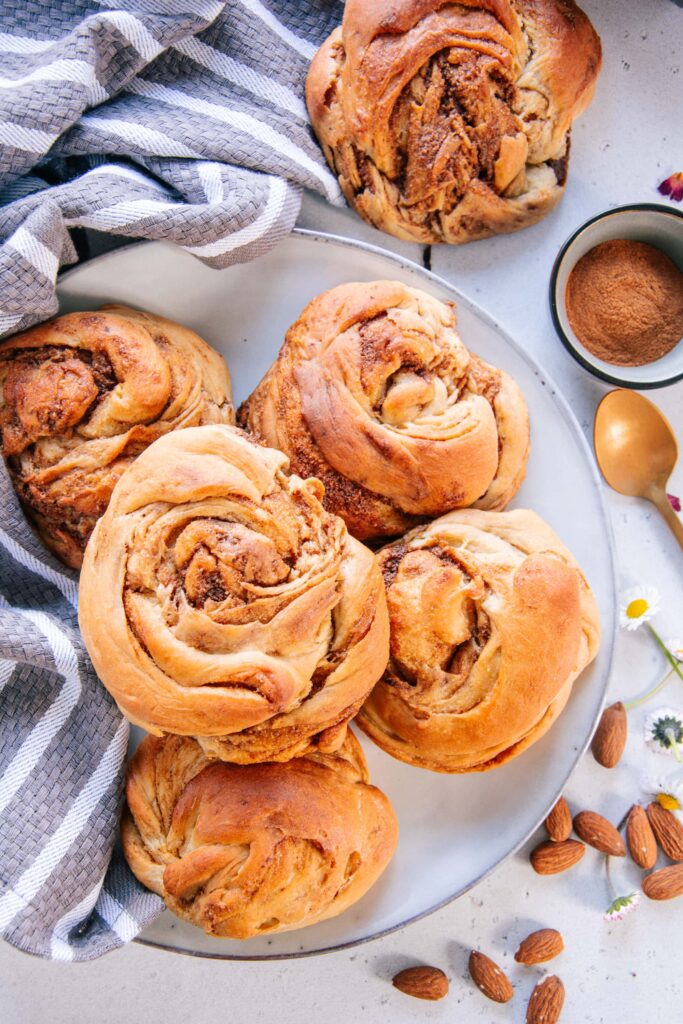 Mandel-Zimtknoten nach dem Grundrezept für veganen süßen Hefeteig ohne Zucker auf einem graublauen Teller mit einem grau-weißen Küchentuch als Dekoration.