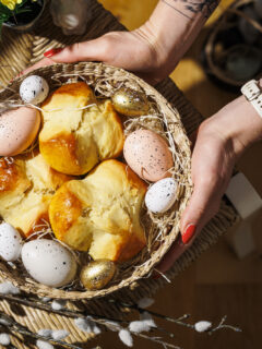 Hände halten Osternest mit bunten Eiern und drei Osterpinzen darin.