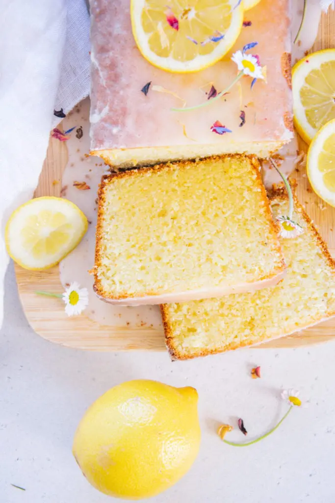 Nahaufnahme von oben von zwei Stücken Zitronenkuchen, die vom ganzen Kuchen auf einem Brett abgeschnitten wurden und neben dem Kuchen aufgefächert liegen. Heruntergelaufene Zitronenglasur auf dem Brett und ganze Zitrone als Verzierung.