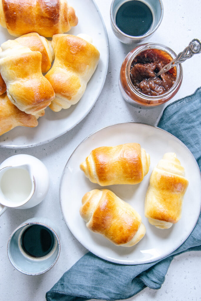 Milchbrötchen auf zwei Teller verteilt und von oben fotografiert. Zwei Espressotassen, eine Milchkanne, ein offenes Marmeladenglas und ein blaues Leinentuch als Dekoration.