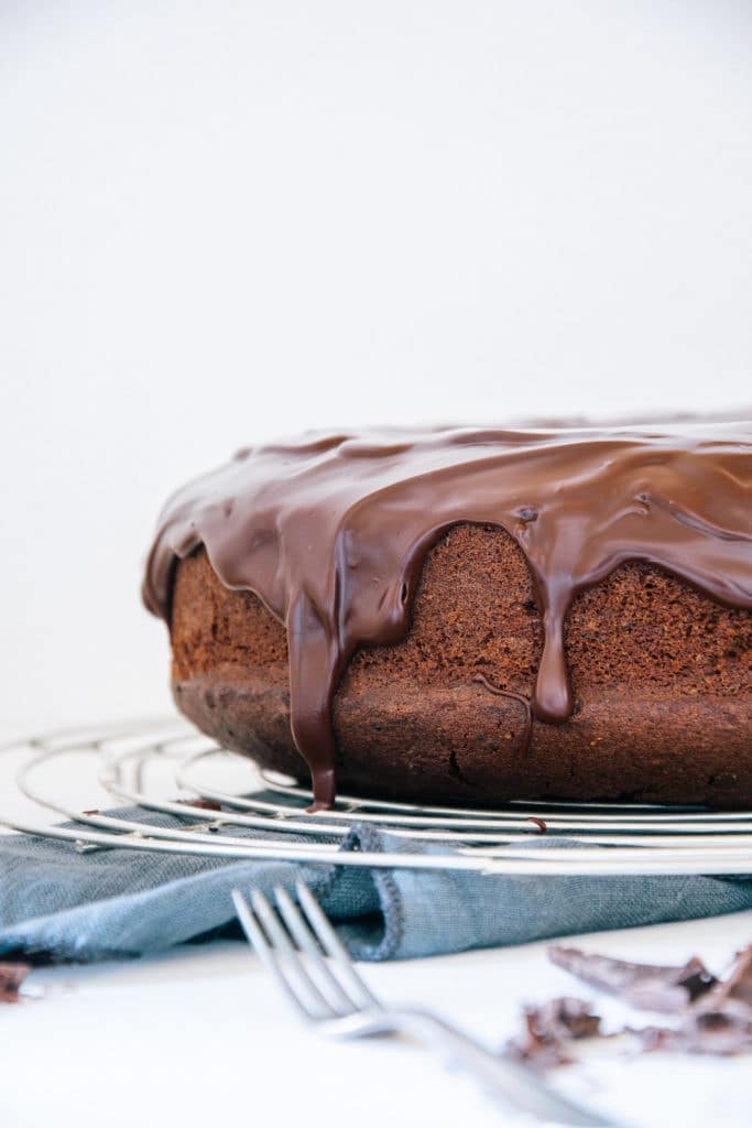 Der beste saftige Schokoladenkuchen auf Augenhöhe von rechts angeschnitten fotografiert, sodass die herabfließende Glasur deutlich erkennbar ist. Kuchen steht auf einem Kuchengitter, darunter ein blaues Leinentuch.