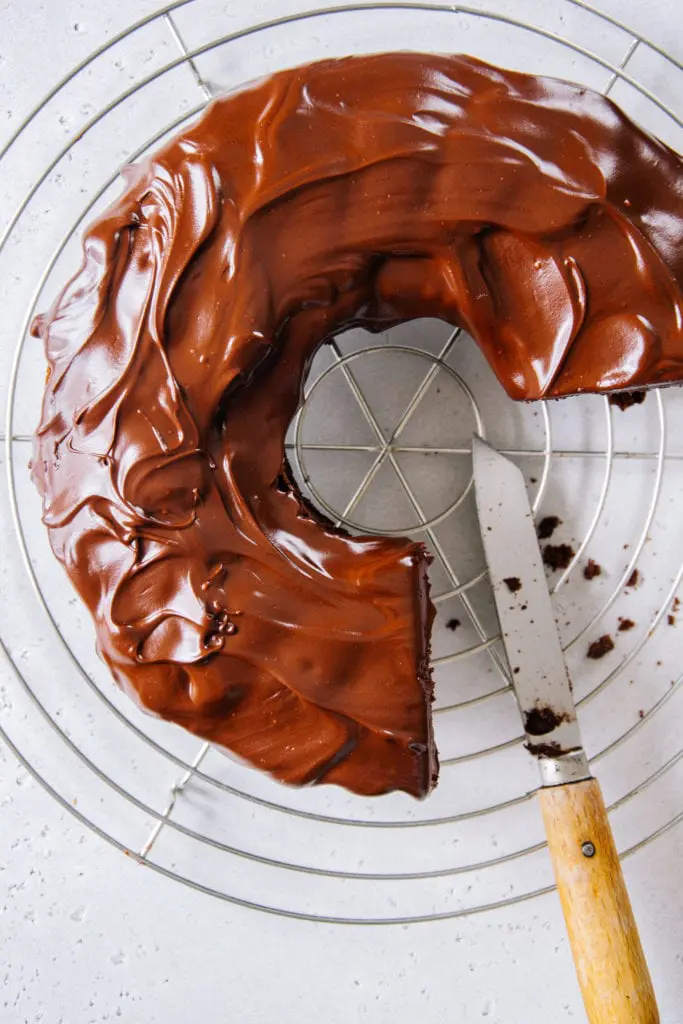 Frisch mit Schokoladenglasur überzogener Schokoladenkuchen auf einem Kuchengitter von oben auf weißem Untergrund fotografiert. Die Glasur ist noch weich und glänzt. Auf dem Kuchengitter liegt ein rustikales Messer mit Holzgriff.