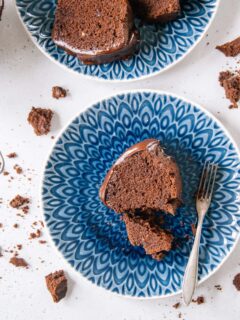 Ein Stück saftiger Schokoladenkuchen von oben auf einem blauen Teller fotografiert. Ein kleiner Teil wurde mit einer alten Kuchengabel abgestochen. Kuchenbrösel auf weißem Untergrund als Verzierung.