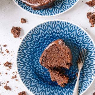 Ein Stück saftiger Schokoladenkuchen von oben auf einem blauen Teller fotografiert. Ein kleiner Teil wurde mit einer alten Kuchengabel abgestochen. Kuchenbrösel auf weißem Untergrund als Verzierung.